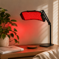 A red light therapy panel lamp on a black adjustable arm, placed on a wooden table next to a potted plant and a book, casting a warm red glow on the wall.